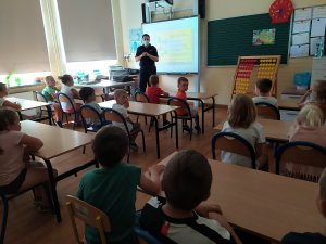 Uczniowie siedzą w ławkach. Odwróceni są plecami. Policjantka stoi przy tablicy interaktywnej i opowiada o zasadach bezpieczeństwa
