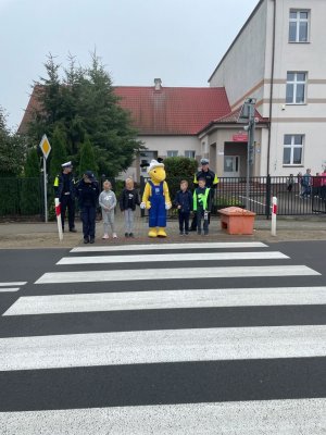 Uczniowie ze Szkoły Podstawowej w Wałdowie stoją przed przejściem dla pieszych. Pomiędzy nimi stoi maskotka Mrówka. Po lewej stronie stoi policjantka. Z tyłu za nimi stoją policjanci ruchu drogowego.