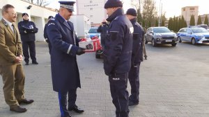 Nadinspektor Piotr Leciejewski wręcza policjantom zakupiony sprzęt fotograficzny. W oddali widać zaparkowane radiowozy policyjne.