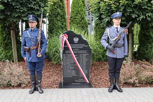 Plac przed komendą Policji w Sępólnie Krajeńskim. Na wprost tablica upamiętniająca pomordowanych policjantów podczas II Wojny Światowej. Po lewej i prawej stronie tablicy stoją policjanci.