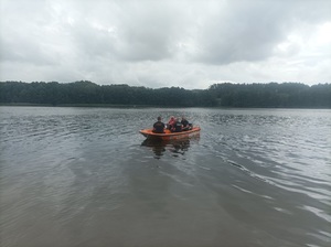 Jezioro Sępoleńskie. Łódź w kolorze pomarańczowym znajduje się na wodzie. W środku siedzi dwóch policjantów, dwóch strażaków oraz dwóch ratowników WOPR.W oddali widać drzewa.