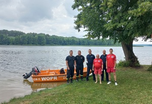 W oddali widać Jezioro Sępoleńskie . Na brzegu do zdjęcia pozują od lewej strony dwóch strażaków, dwóch policjantów i dwóch ratowników WOPR. Po prawej strony widać duże drzewo. Z tyłu za nimi stoi przycumowana pomarańczowa łódź.