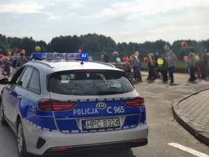 Przed stadionem miejskim w kamieniu Krajeńskim stoi radiowóz policyjny. Za nim w oddali widać uczestników marszu