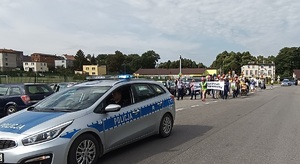 Trasa przemarszu przy stadionie miejskim w kamieniu krajeńskim. Z przodu jedzie policyjny radiowóz, a za nim idą uczestnicy marszu z transparentami.