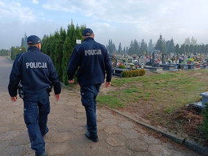 Dwóch umundurowanych policjantów patroluje okolice cmentarza. Po prawej stronie widać groby zmarłych.