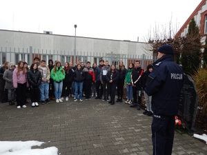 Skwer przy komendzie . Komendant inspektor Paweł Zawada opowiada o historii Policji. Przed nim stoi ustawiona w rzędzie młodzież. W tle widać budynki komendy.