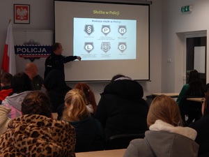 Świetlica w komendzie . Uczniowie klas policyjnych siedzą w ławkach. Przed nimi stoi policjant, który omawia stopnie policyjne. W tle prezentacja multimedialna