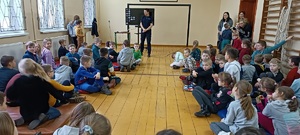 Sala gimnastyczna w szkole. Uczniowie zwróceni są plecami do zdjęcia. Siedza na materacach. przed nimi stoi policjantka, a za nią na tablicy wyświetla się prezentacja multimedialna.