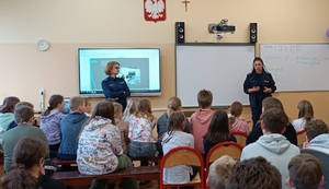 Pomieszczenie sali lekcyjnej. Policjantki stoją przed uczniami, Zwrócone są twarzami do zdjęcia. Przed nimi w łąkach szkolnych siedzą uczniowie. Za policjantkami, na ścianie wyświetla się prezentacja multimedialna.