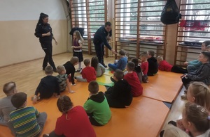 Sala gimnastyczna w szkole. Policjanci stoją, dzieci siedzą na materacach. Zwróceni są plecami do zdjęcia. Funkcjonariusze rozdają dzieciom odblaski.