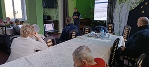 Świetlica w Domu Pomocy Społecznej. Pensjonariusze siedzą przy stołach razem z dzielnicowymi. zwróceni sa plecami do zdjęcia. W oddali widać stojącą przed nimi policjantkę, a za nią na tablicy wyświetla się prezentacja. Policjantka zwrócona jest twarzą do zdjęcia.