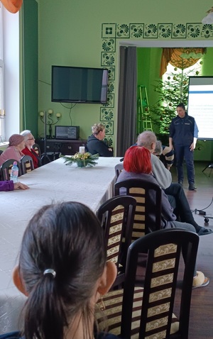 Świetlica w Domu Pomocy Społecznej w Kamieniu Krajeńskim. Po lewej stronie zdjęcia widać   pensjonariuszy którzy siedzą na krzesłach przy stołach. Zwróceni są plecami do zdjęcia. W oddali widać stojącą przed nimi policjantkę, a za nią na tablicy wyświetla się prezentacja. Policjantka zwrócona jest twarzą do zdjęcia.