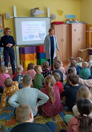 Sala w przedszkolu w Jastrzębcu. Policjantka razem z przedstawicielem sanepidu stoją przed dziećmi. Przedszkolaki siedzą przed nimi  na podłodze. Za plecami gości wyświetla się prezentacja multimedialna dotycząca bezpiecznych ferii.
