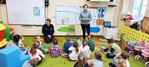 Sala w przedszkolu w Przepałkowie. Policjantka razem z dziećmi siedzi na podłodze. Przedstawiciel sanepidu stoją przed dziećmi i opowiada o zdrowym spędzaniu czasu wolnego.  Za plecami gości wyświetla się prezentacja multimedialna dotycząca bezpiecznych ferii.