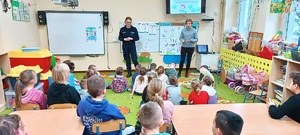 Sala w przedszkolu w Przepałkowie. Policjantka razem z przedstawicielem sanepidu stoją przed dziećmi. Przedszkolaki siedzą przed nimi  na podłodze. Za plecami gości wyświetla się prezentacja multimedialna dotycząca bezpiecznych ferii.