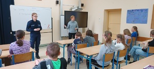 Klasa szkolna w Przepałkowie. Policjantka razem z przedstawicielem sanepidu stoją przed uczniami. Zwrócone są twarzami do zdjęcia. Przed nimi w ławkach szkolnych siedzą uczniowie. Za plecami gości wyświetla się prezentacja multimedialna dotycząca bezpiecznych ferii.