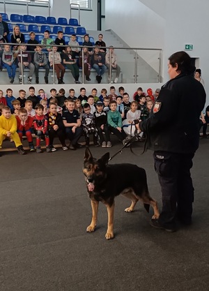 Sala gimnastyczna w Szkole Podstawowej nr 1 w Więcborku. Uczniowie siedzą na trybunach. Policjantka razem z psem oraz przedstawicielem sanepidu stoją przed dziećmi.
