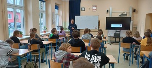 Pomieszczeni klasy. Uczniowie siedzą w ławkach szkolnych. Nie widać ich twarzy. Przed nimi stoi policjantka oraz przedstawiciel sanepidu. Z ich plecami znajduje się tablica szkolna.