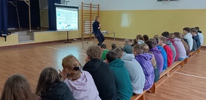 Sala gimnastyczna w Jastrzębcu. Uczniowie siedzą na ławkach. Zwróceni są do zdjęcia plecami. Przed nimi stoi policjantka . Za jej plecami wyświetla się prezentacja multimedialna.