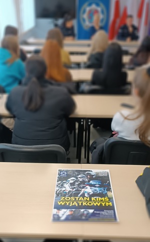 Świetlica w Komendzie Powiatowej policji w Sępólnie Krajeńskim. W tle uczniowie którzy siedzą w ławkach, plecami do zdjęcia. Na stole leży plakat z napisem :  zostań kimś wyjątkowym,