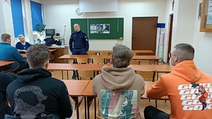 Klasa lekcyjna w Zespole Szkół Ponadpodstawowych w Sępólnie Krajeńskim . Uczniowie siedzą w ławkach szkolnych. Przed nimi stoi policjant . W tle widać zawieszone na tablicy plakaty promujące zawód policjanta.