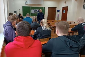 Klasa lekcyjna w Zespole Szkół Ponadpodstawowych w Sępólnie Krajeńskim. Uczniowie siedzą w ławkach szkolnych. Przed nimi stoi policjant . W tle widać zawieszone na tablicy plakaty promujące zawód policjanta.