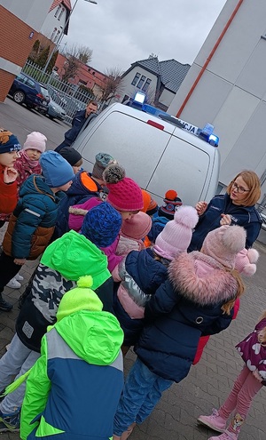 Plac przy Komendzie Powiatowej Policji w Sępólnie Krajeńskim. Dzieci stoją przy radiowozie policyjnym, zwrócone są plecami do zdjęcia. Przed nimi stoi policjantka, która pokazuje im policyjny radiowóz. W oddali widać budynki.