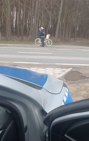 Zdjęcie wykonane zza radiowozu. W oddali, po drugiej stronie jezdni widać policjanta, który kontroluje zatrzymanego rowerzystę.