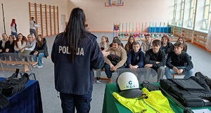 Sala gimnastyczna w Liceum Ogólnokształcącym w Sępólnie Krajeńskie. Policjantka stoi plecami do zdjęcia. Przed nią na ławkach siedzą uczniowie. Po prawej stronie zdjęcia widać rozłożony na stoisku sprzęt policyjny.