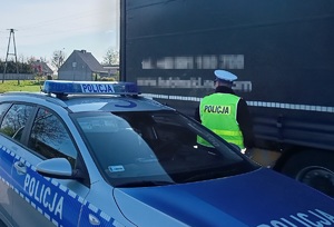Policjant stoi plecami do zdjęcia. Przed nim znajduje się naczepa pojazdu ciężarowego, a za jego plecami policyjny radiowóz