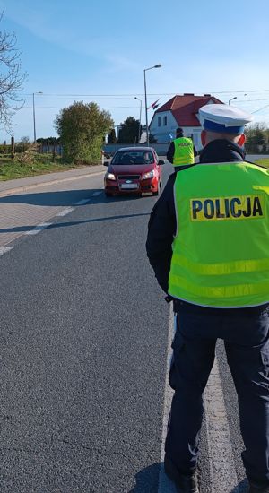 Policjant stoi na ulicy i kontroluje pojazdy. W oddali widać budynki