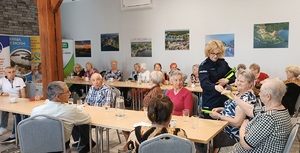 Policjantka rozdaje ulotki uczestnikom spotkania. Seniorzy siedzą przy stolikach, pomiędzy nimi stoi policjantka