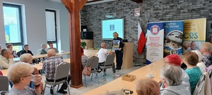 Sala konferencyjna w Centrum Aktywności Społecznej w Więcborku. Policjantka stoi przed seniorami, którzy siedzą przy stołach. Za plecami policjantki wyświetla się prezentacja multimedialna z napisem moja komenda.