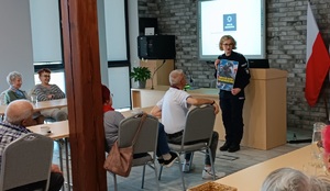 Sala konferencyjna w Centrum Aktywności Społecznej w Więcborku. Policjantka stoi przed seniorami, którzy siedzą przy stołach. Za plecami policjantki wyświetla się prezentacja multimedialna z napisem moja komenda. Funkcjonariuszka trzyma w rękach plakat promujący zawód policjanta.
