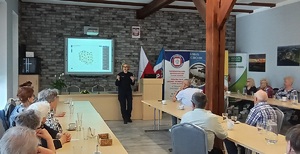 Sala konferencyjna w Centrum Aktywności Społecznej w Więcborku. Policjantka stoi przed seniorami, którzy siedzą przy stołach. Za plecami policjantki wyświetla się prezentacja multimedialna z Krajowa Mapą zagrożenia Bezpieczeństwa.