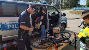 Festyn z okazji Dnia Dziecka nad Jeziorem Mochel w Kamieniu Krajeńskim. Policjantka znakuje ułożony na ławce rower. Koło niej stoi drugi policjant. Za policjantami widać zaparkowany radiowóz. Przy radiowozie stoi uczestnik festynu.