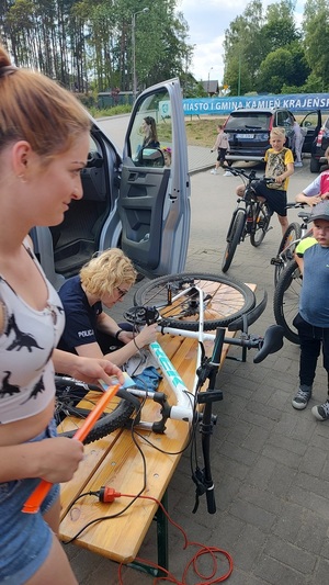 Festyn z okazji Dnia Dziecka nad Jeziorem Mochel w Kamieniu Krajeńskim. Policjantka znakuje ułożony na ławce rower. Koło niej znajduje się kolejka rowerów do znakowania. Za policjantka widać zaparkowany radiowóz. Przy radiowozie stoi uczestnik festynu.