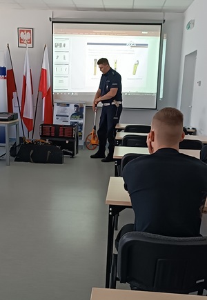 Uczniowie klasy policyjnej siedzą w ławkach. Przed nimi stoi policjant ruchu drogowego i opowiada o swojej pracy. Pokazuje przedmioty służące do wykonywania pomiarów na miejscu wypadku drogowego.