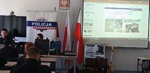 Uczniowie klasy policyjnej siedzą w ławkach. Przed nimi stoi policjant ruchu drogowego i opowiada o swojej pracy. W tle wyświetla się prezentacja multimedialna.