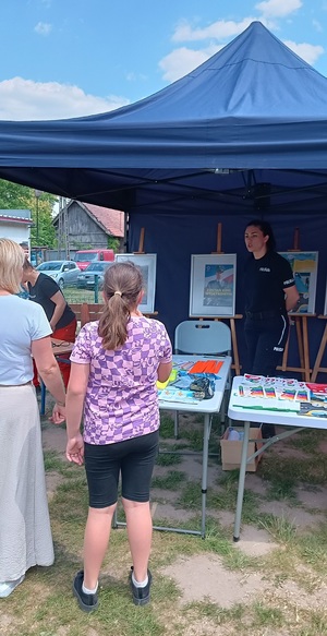 Policjantka rozmawia z uczestnikami festynu. Stoi pod namiotem, a przed nią na stole leża ulotki i odblaskowe kamizelki.