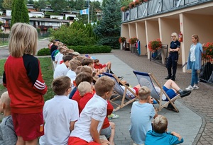 Policjantka wspólnie z pracownikiem sanepidu staja przed dziećmi. W tle widać budynek ośrodka wczasowego. Dzieci siedzą na murku i krzesłach.