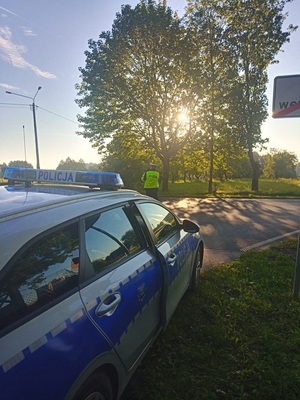 Radiowóz policyjny stoi przy drodze. Za nim widać policjanta ruchu drogowego, który stoi przy drodze i dokonuje pomiaru prędkość pojazdów.