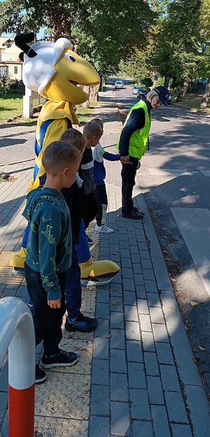 Policjantka uczy dzieci zasad bezpiecznego poruszania się w drodze do szkoły. Dzieci razem z policjantką i maskotką Mrówki stoją przed przejściem dla pieszych.