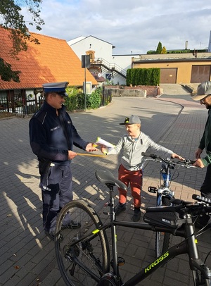 Policjant wręcza chłopcu z rowerem kamizelkę odblaskową. W oddali widać pobliskie zabudowania.