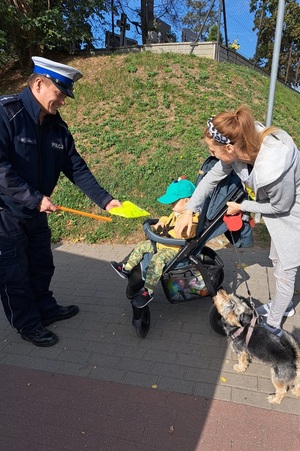 Policjant wręcza dziecku siedzącemu w wózku kamizelkę odblaskową. Obok wózka dziecięcego stoi kobieta.