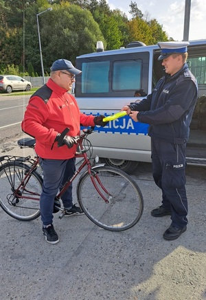 Policjant wręcza zatrzymanemu rowerzyście kamizelkę odblaskową. W tle widać policyjny radiowóz.