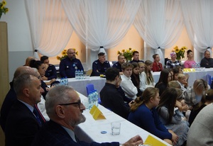 Debata w Lutowie. Sala lekcyjna. Zaproszeni goście siedzą przy stolikach. Pośród gości siedzą policjanci. Przed nimi na ławeczkach szkolnych siedzą uczniowie.