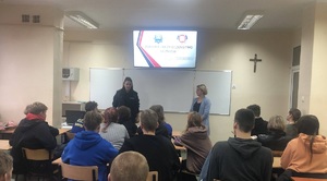 Policjantka wspólnie z przedstawicielem sanepidu stoją przed uczniami którzy siedzą w ławkach szkolnych. W tle widać prezentacja multimedialną z napisem &quot;Zdrowie i bezpieczeństwo uczniów&quot;.