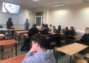 Policjantka wspólnie z przedstawicielem sanepidu stoją przed uczniami którzy siedzą w ławkach szkolnych. W tle widać prezentacja multimedialną z napisem &quot;Zdrowie i bezpieczeństwo uczniów&quot;.