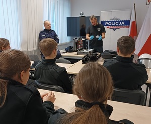 Uczniowie siedzą przy stolikach. przed nimi stoją policjanci opowiadający o swojej służbie.